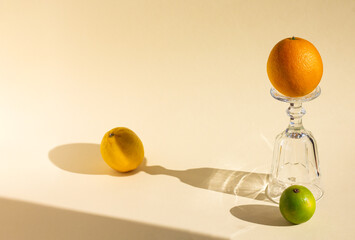 art creative composition of upside down empty goblet, orange, lemon and lime, crystal glass and fruits are under hard light on soft beige background, long glitter harsh shadows, high angle