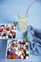 Greek salad in square plates and lemonade on a light blue background.
