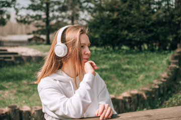 Naklejka premium A girl in a white hoodie listens to music with headphones and relaxes sitting on a bench near a country house in nature, ASMR concept