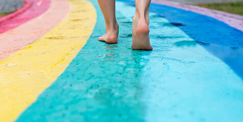 Female feet close up on barefoot on wet asphalt drawn rainbow walking path after rain among...