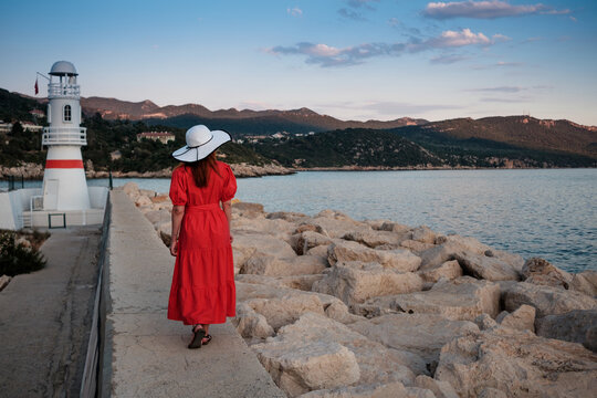 Beautiful picture of a  girl in a hat and red dress near the lighthouse and boats  at sunset. Port of Kas old town, Antalya, Turkey.