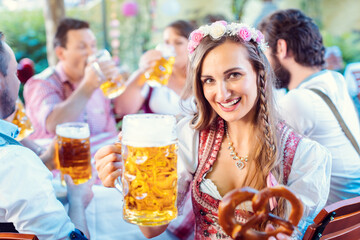 Woman toasting to the camera with glass of beer in Bavarian pub holding pretzel in hand
