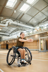 Vertical full length portrait of young woman in wheelchair warming up before sports practice at volleyball court