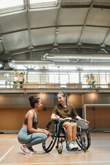 Fototapeta na wymiar Vertical full length portrait of young woman in wheelchair talking to coach or teammate during sports practice at indoor court
