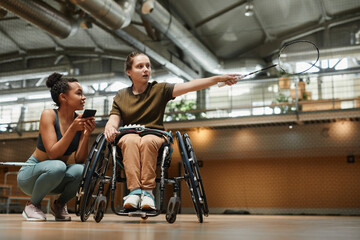 Fototapeta na wymiar Full length portrait young woman in wheelchair talking to coach during badminton practice at sports court, copy space