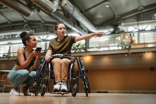 Full length portrait young woman in wheelchair talking to coach during badminton practice at sports court, copy space