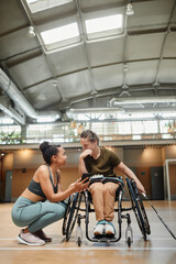 Vertical full length portrait young woman in wheelchair talking to friend during badminton practice at sports court