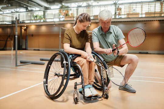 Full length portrait of smiling young woman in wheelchair talking to coach during badminton practice at sports court