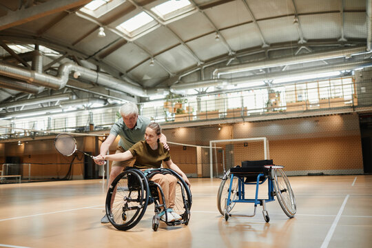 Full length portrait of mature coach training young woman in wheelchair during badminton practice in sports court, copy space