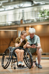 Full length portrait of senior coach training young woman in wheelchair during badminton practice in sports court and using smartphone