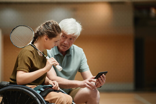 Side view portrait of senior coach training young woman in wheelchair during badminton practice in sports court and using smartphone, copy space