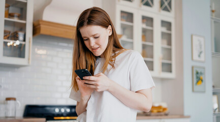 A freelance blogger texts a friend on the phone. Red-haired female appearance, lunch break at home. He smiles and looks at the screen of the gadget. Stylish kitchen in the apartment