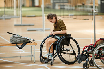 Full length portrait of young woman strapping wheelchair while getting ready for sports practice at indoor court, copy space