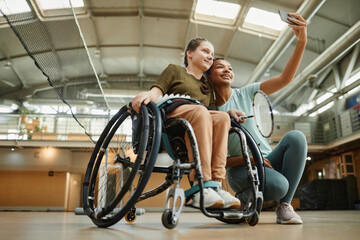 Low angle portrait of young woman in wheelchair taking selfie with friend at indoor sports court, copy space - Powered by Adobe