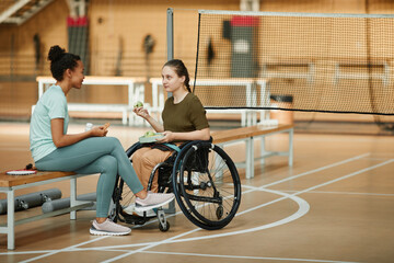 Full length portrait of young woman in wheelchair sharing healthy snack with friend at indoor sports court, copy space