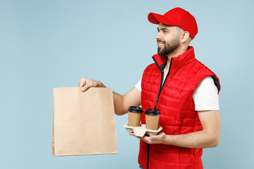 Delivery guy employee man in red cap white T-shirt vest uniform work courier service hold brown clear blank craft paper takeaway bag cup coffee to go isolated on pastel blue color background studio - Powered by Adobe