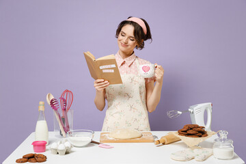 Young happy housewife housekeeper cook chef baker woman in pink apron work at table kitchenware hold cup coffee recipe book drink tea isolated on pastel violet background Process cooking food concept.