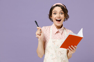 Young housewife housekeeper chef cook baker woman in pink apron write down recipe in cook book notebook point finger up with new idea isolated on pastel violet background Cooking food process concept