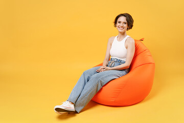 Full length young smiling happy friendly woman 20s with bob haircut wearing white tank top shirt sit in orange bag chair isolated on yellow color background studio portrait. People lifestyle concept