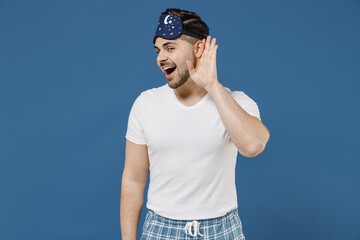 Young curious nosy man 20s wear pajamas jam sleep mask white t-shirt rest relax at home try to hear...