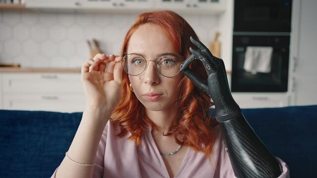 Young red-haired attractive woman putting on glasses using bionic prosthesis. The girl is adjusting the bionic prosthesis. Bionics Cybernetic Robotic arm