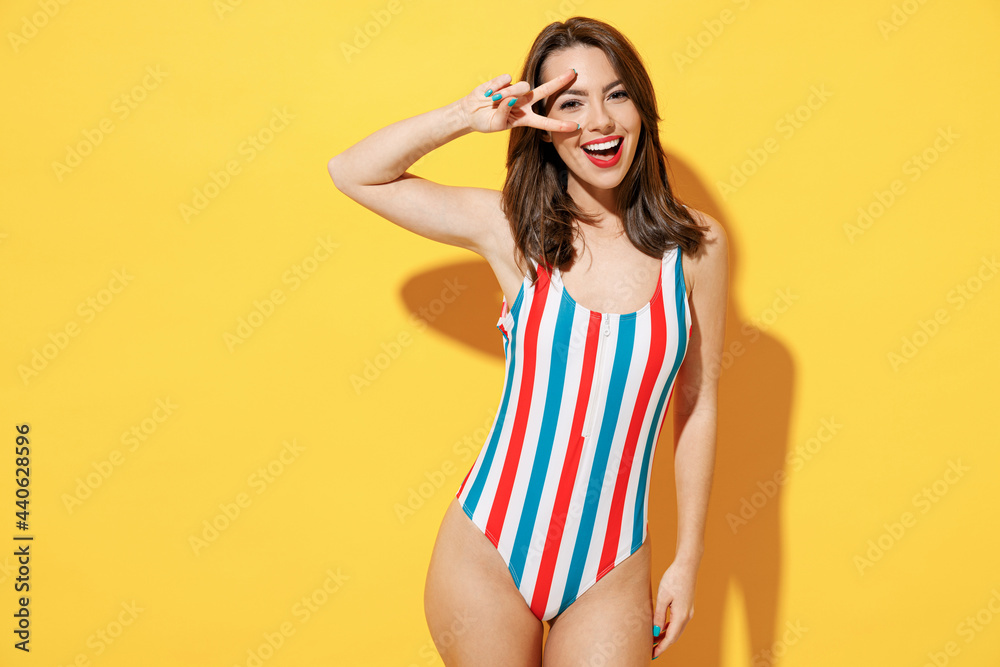 Wall mural Happy young sexy woman slim body wear red blue one-piece swimsuit showing victory sign v-sign gesture isolated on vivid yellow color wall background studio. Summer hotel pool sea rest sun tan concept.