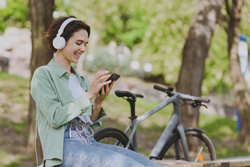 Obraz premium Young woman in casual green jacket jeans headphones sit on bench near bicycle bike in city spring park outdoors hold mobile cell phone listen to music chat online People active urban youthful concept.