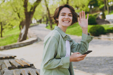 Young smiling friendly woman 20s in casual green jacket jeans sitting on bench in city spring park...
