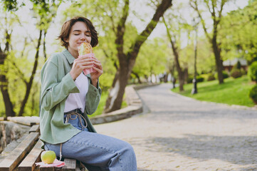 Young student woman 20s in casual green jacket jeans sitting on bench in city spring park outdoors...