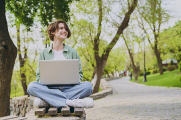 Full length young student freelancer woman in green jacket jeans sit on bench in spring park...