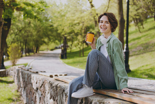 Young smiling fun woman in casual green jacket jeans sit on bench in city spring park outdoors hold takeaway delivery craft paper brown cup coffee to go, rest in morning People urban youthful concept.