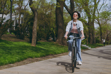 Full length young sporty woman 20s wear casual jeans clothes headphones riding bicycle bike on sidewalk in city spring park outdoors, look aside. People active urban healthy lifestyle cycling concept