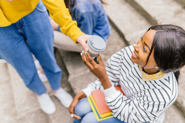 Smiling african female student sitting on the stairs take cup of coffee from unrecognizable woman....