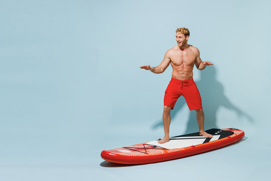 Full length young sexy sporty strong man 20s wearing red shorts swimsuit relax stand on orange sup board surfing training isolated on pastel blue background. Summer vacation sea rest sun tan concept