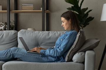 Side view happy young woman sitting on couch with laptop on laps, working distantly on project, using software applications, communicating distantly or web surfing information online alone at home.