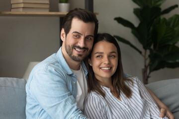 Portrait of sincere happy dreamy young family couple cuddling, resting on comfortable sofa. Affectionate loving millennial man and woman looking in distance, imagining planning future at home.