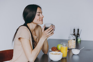 Side view young housewife woman 20s wear casual clothes beige shirt eat breakfast hold cup of tea...