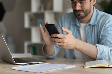 Cropped image distracted from work on laptop young employee businessman using smartphone applications, enjoying break pause time playing online game or shopping, daily modern tech usage concept.