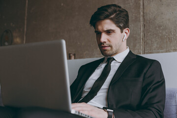 Bottom view serious young employee business man corporate lawyer in formal black suit shirt tie air pods sit on grey sofa in livingroom indoors use laptop pc computer People career lifestyle concept