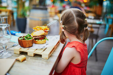 Adorable three year old girl sitting eating hamburger in cafe or restaurant