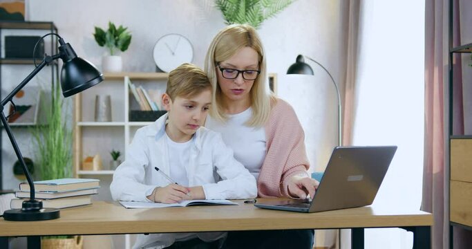 Portrait of attractive skilled caring adult blond mother in glasses which sitting at home desk with her handsome son and helping him with online test ,home education concept