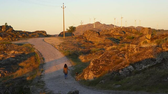 Woman walking with dog on a road in nature mountain landscape with wind turbines at golden hour in Sortelha, Portugal