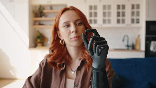 Young red-haired woman looks at her bionic prosthetic left hand and alternately flexes and unbends her fingers. Girl is adjusting the bionic prosthesis Bionics Cybernetic Robotic-arm Hand prosthesis