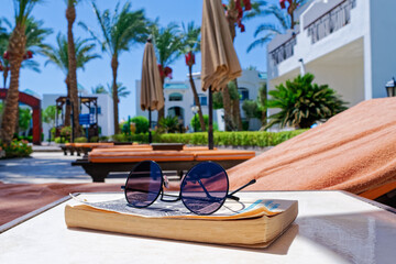 Close-up view of sunglasses on a closed book and sun loungers by the pool. Behind there are sun umbrellas, resort buildings, garden. Vacation concept, reading outdoors, recreation by the sea.