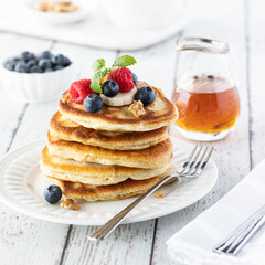 Straight on view of a stack of buttermilk pancakes topped with berries and bananas with syrup in behind.