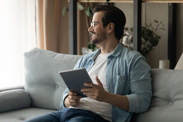 Happy dreamy young man in eyeglasses sitting on comfortable couch with digital computer tablet in hands, feeling inspired of getting message in social networks with good news, modern technology usage.