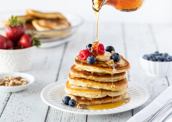 A stack of buttermilk pancakes served with berries and bananas with syrup being poured on top.