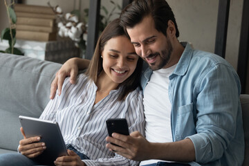 Addicted to modern technology happy young family couple using different gadgets, involved in chatting in social networks, shopping in internet store, spending time online resting together at home.