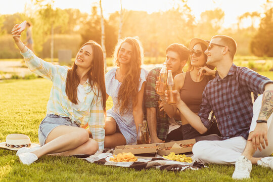 group of young people sitting on a picnic blanket having fun together with pizza and drinks and taking a selfie
