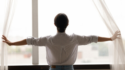 Back view of Indian woman open curtains meet welcome new sunny day in cozy home. Mixed race...